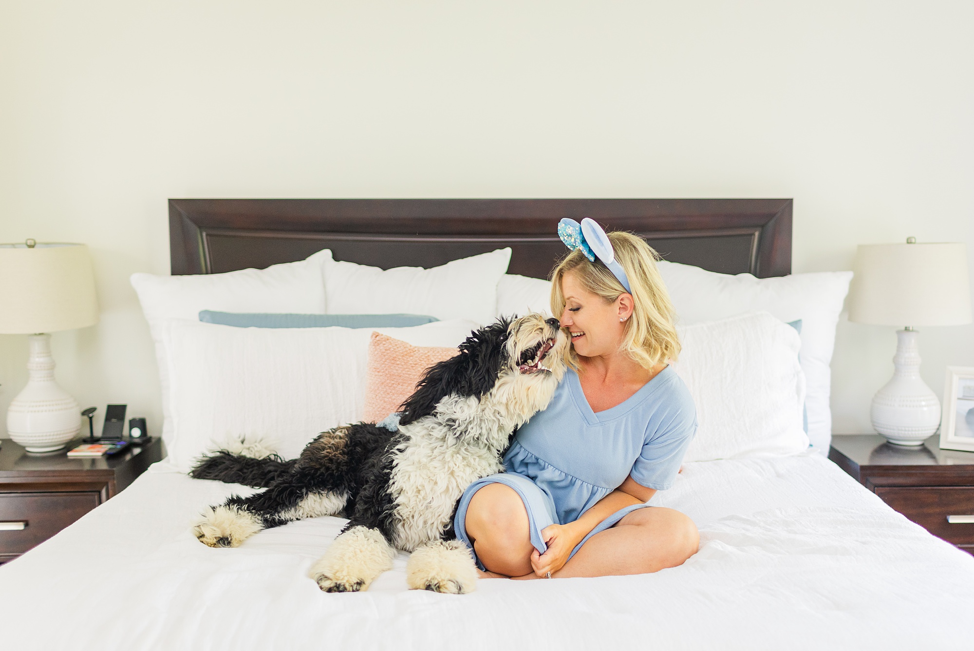 woman laughs with dog on bed wearing blue dress and Mickey Ears