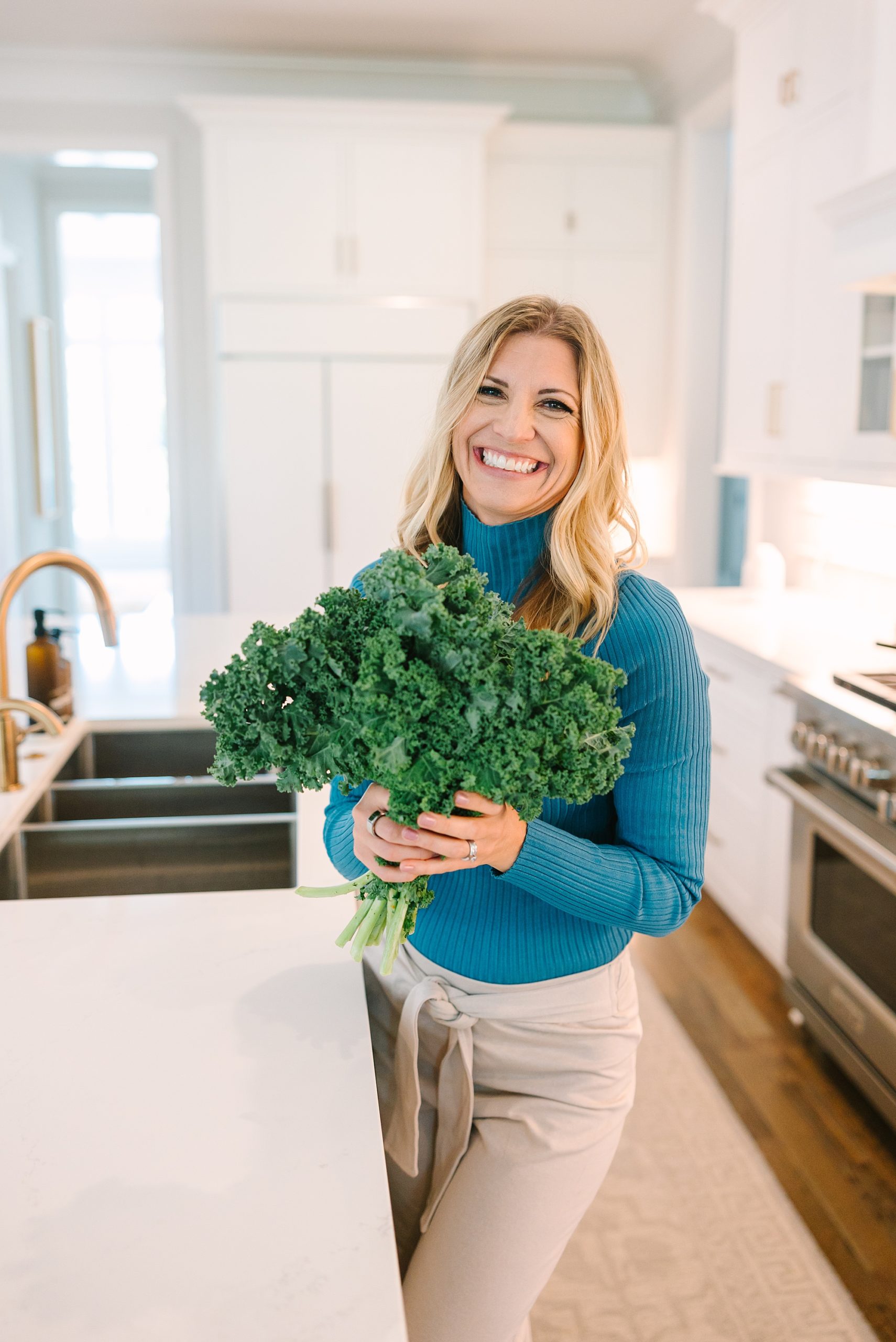 woman in teal turtleneck stands in kitchen holding kale