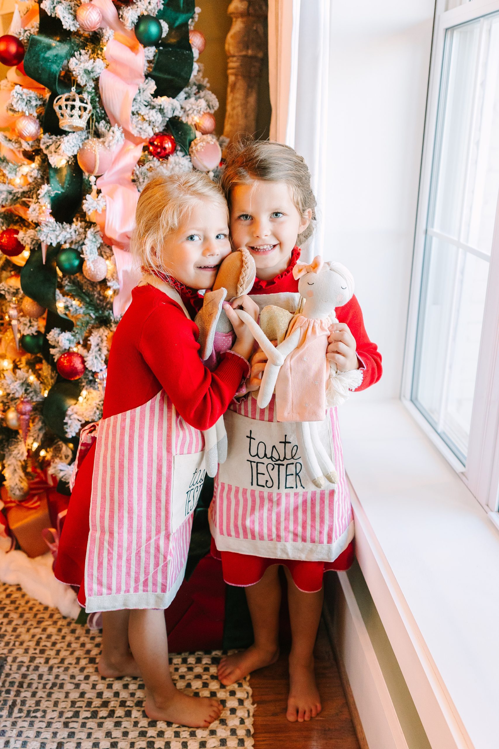 girls smile by window hugging dolls in Christmas aprons