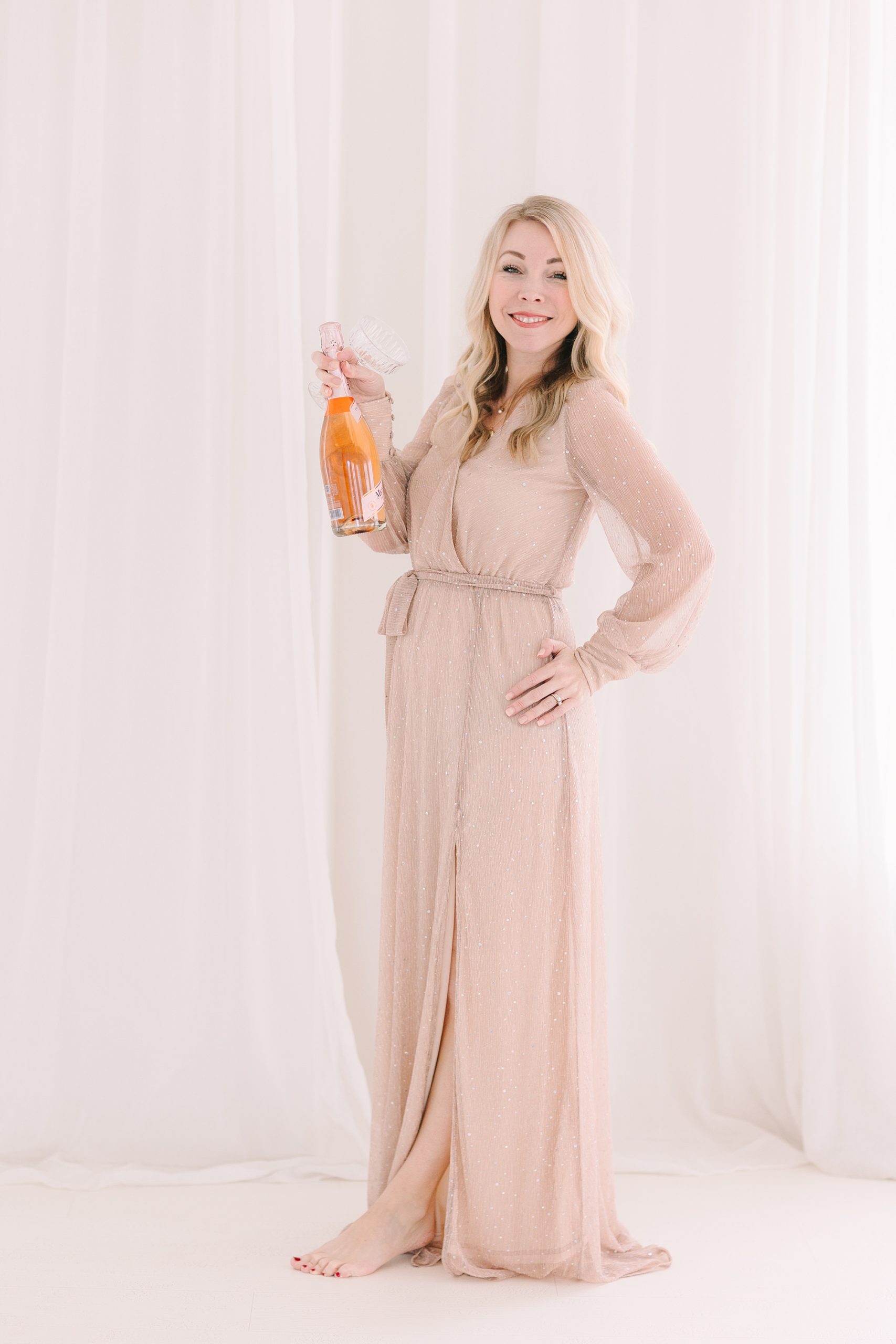 woman in floor-length tan dress holds bottle of pink champagne