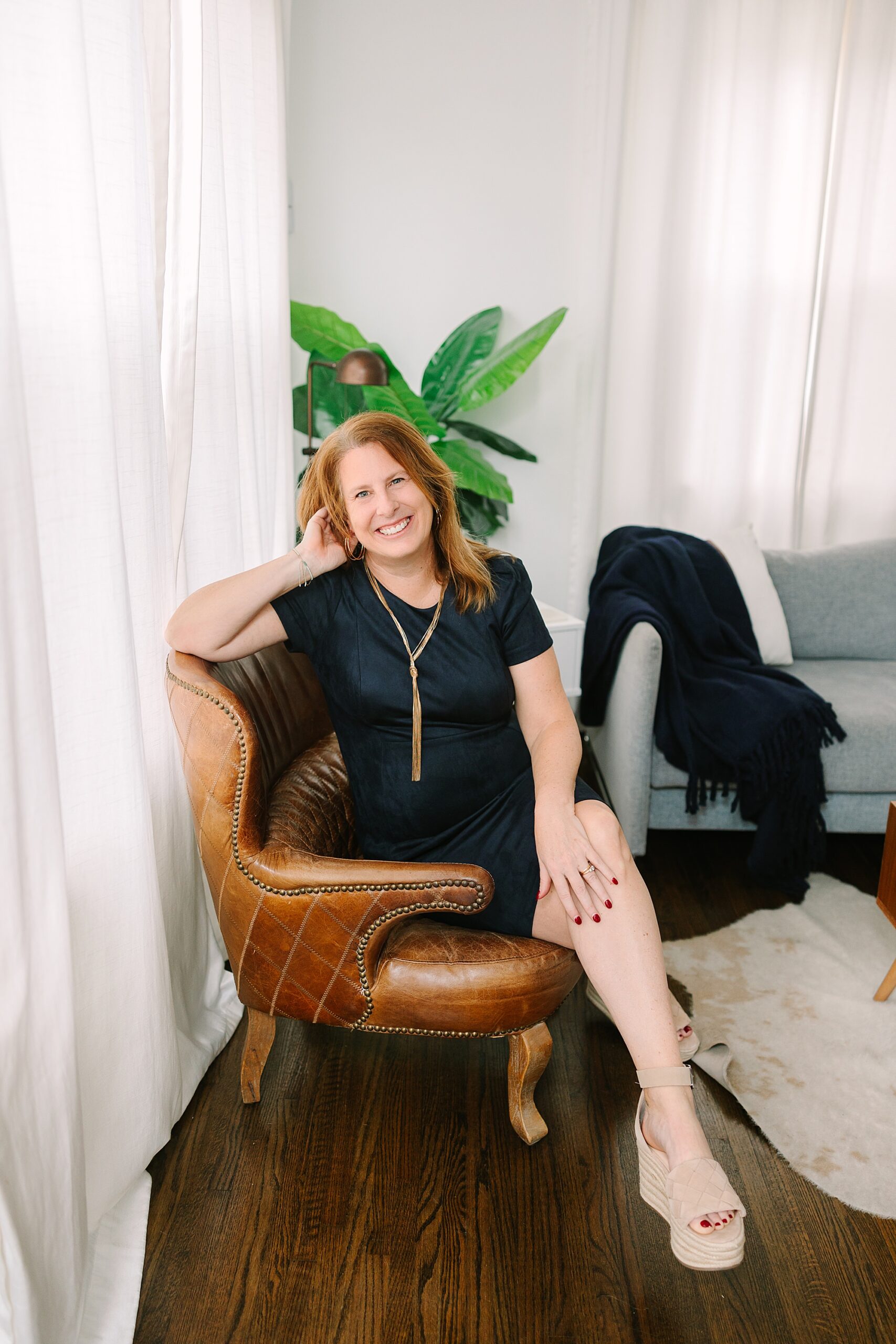 woman sits in leather chair wearing black dress during branding photos