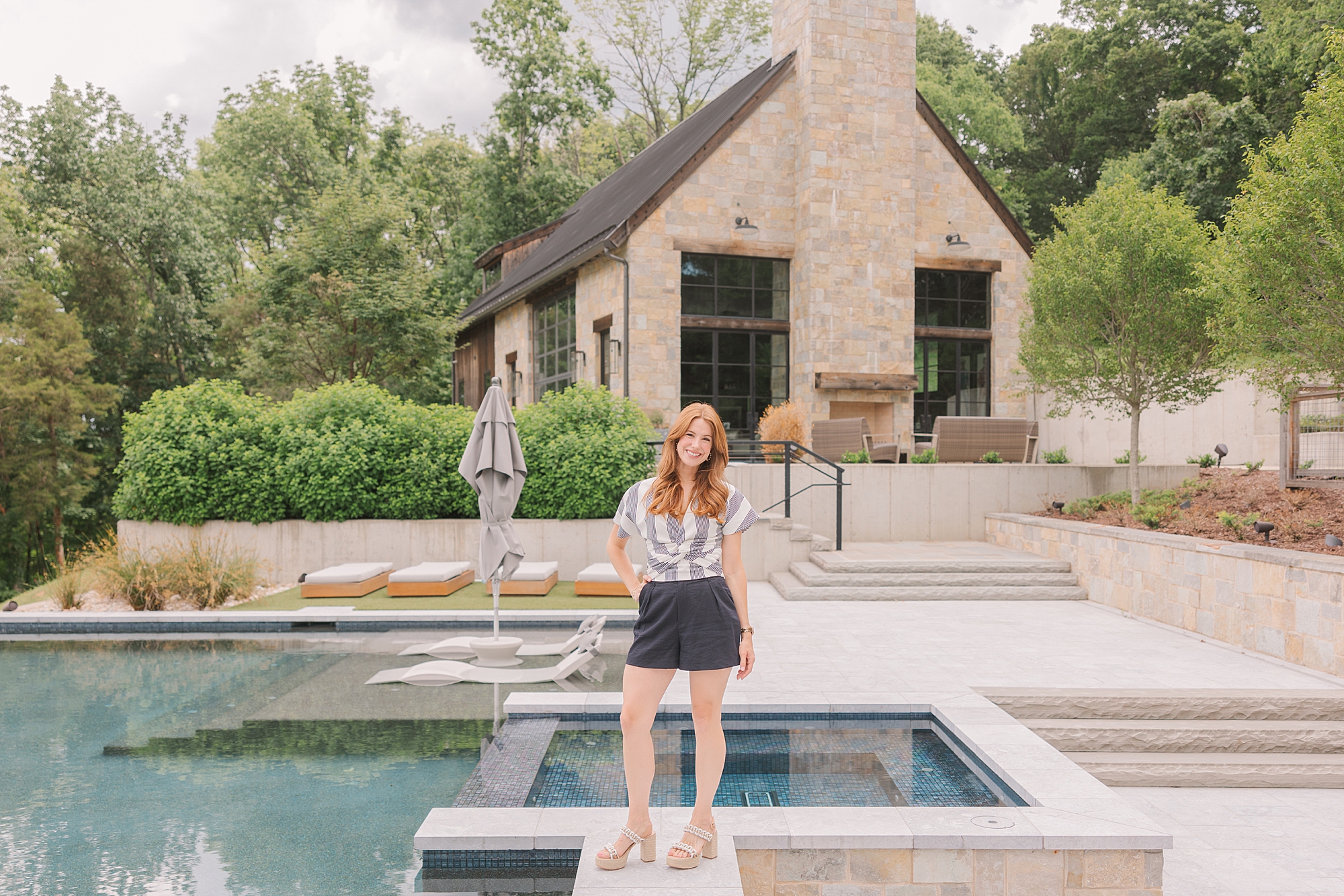 redhead woman stands by pool in striped grey shirt with skirt during branding session