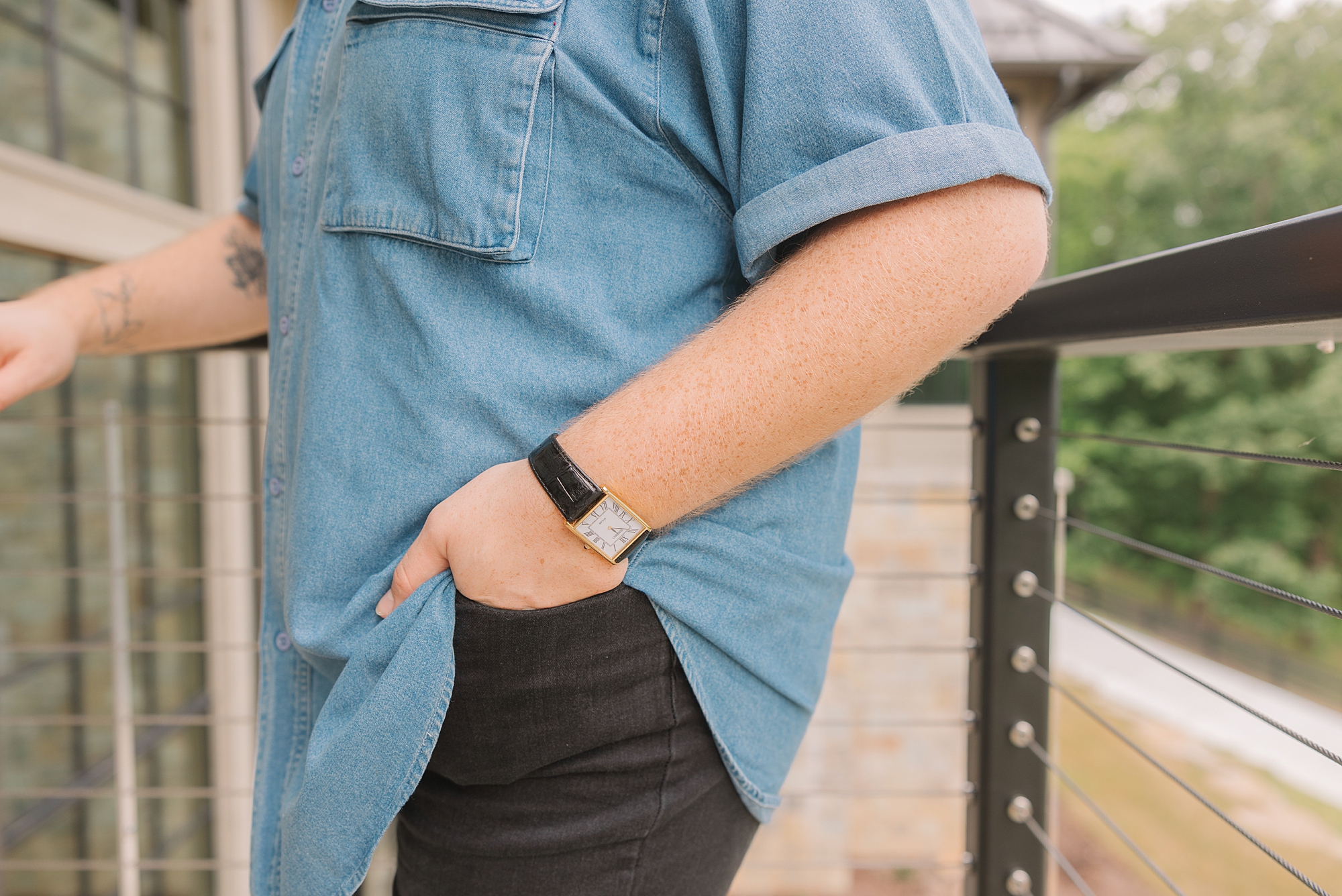man in blue shirt stands with hand in pocket showing off watch