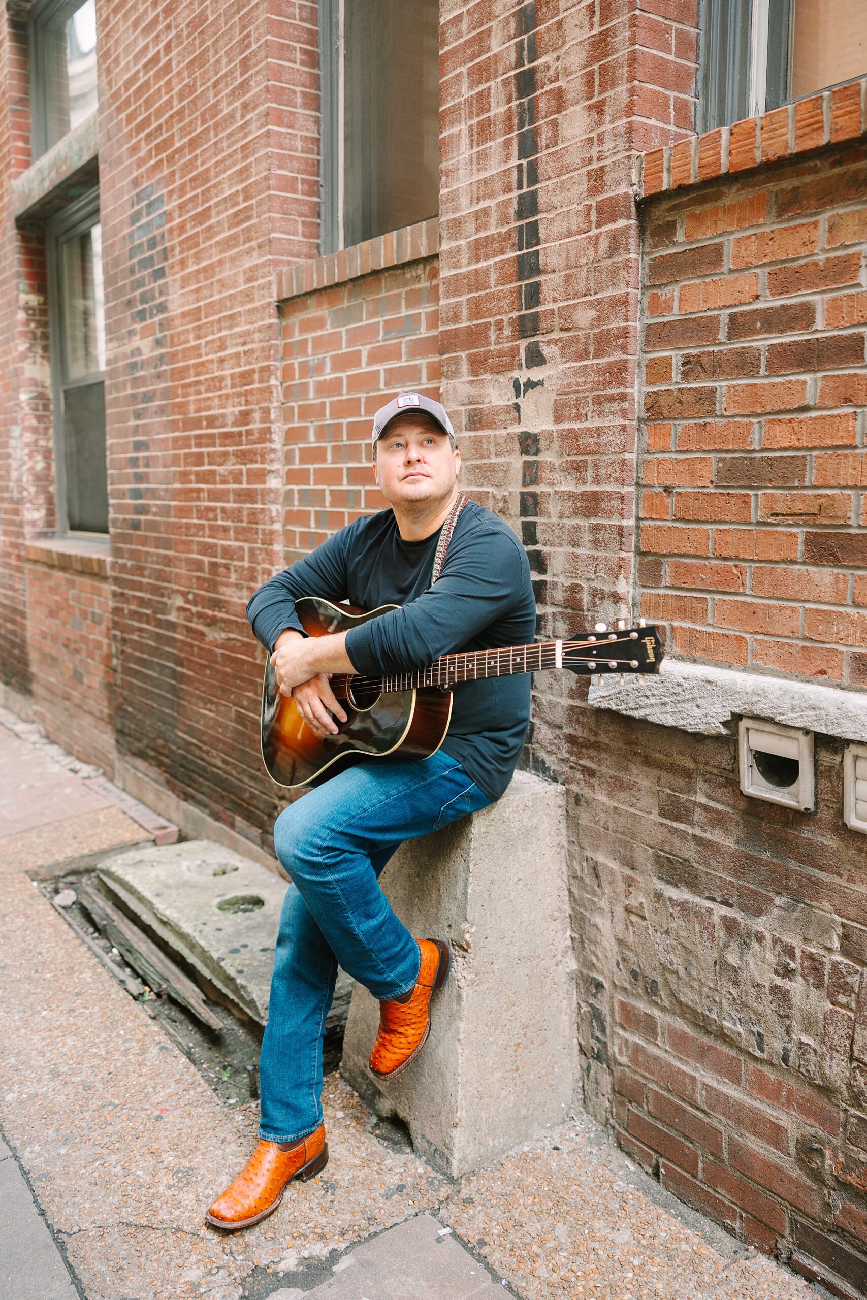 singer/songwriter leans against brick wall holding guitar against him in Downtown Nashville