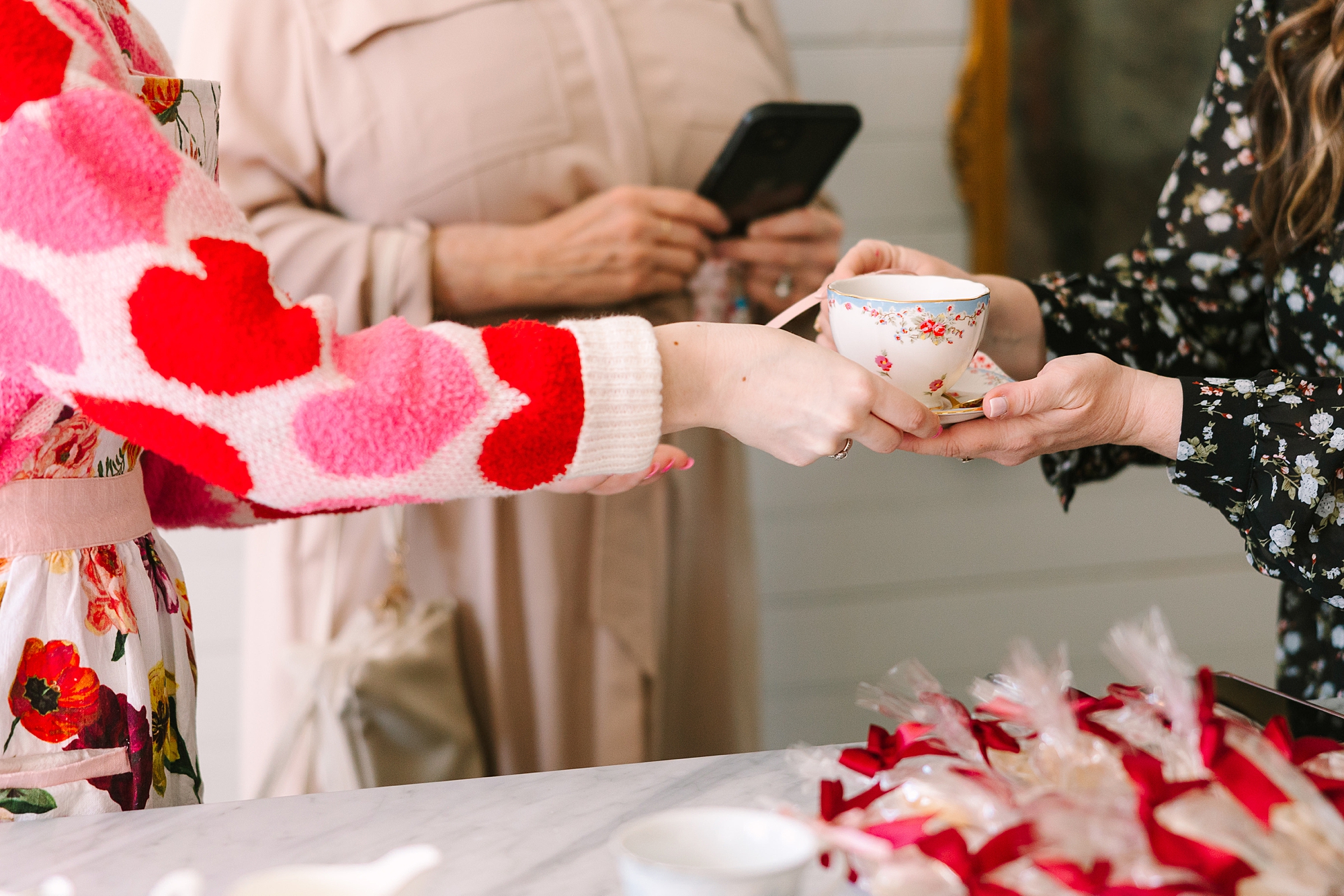 woman in sweater with red and pink hearts hands tea cup to friend during Galentine's Day Tea hosted by Pretty Lovely Teas