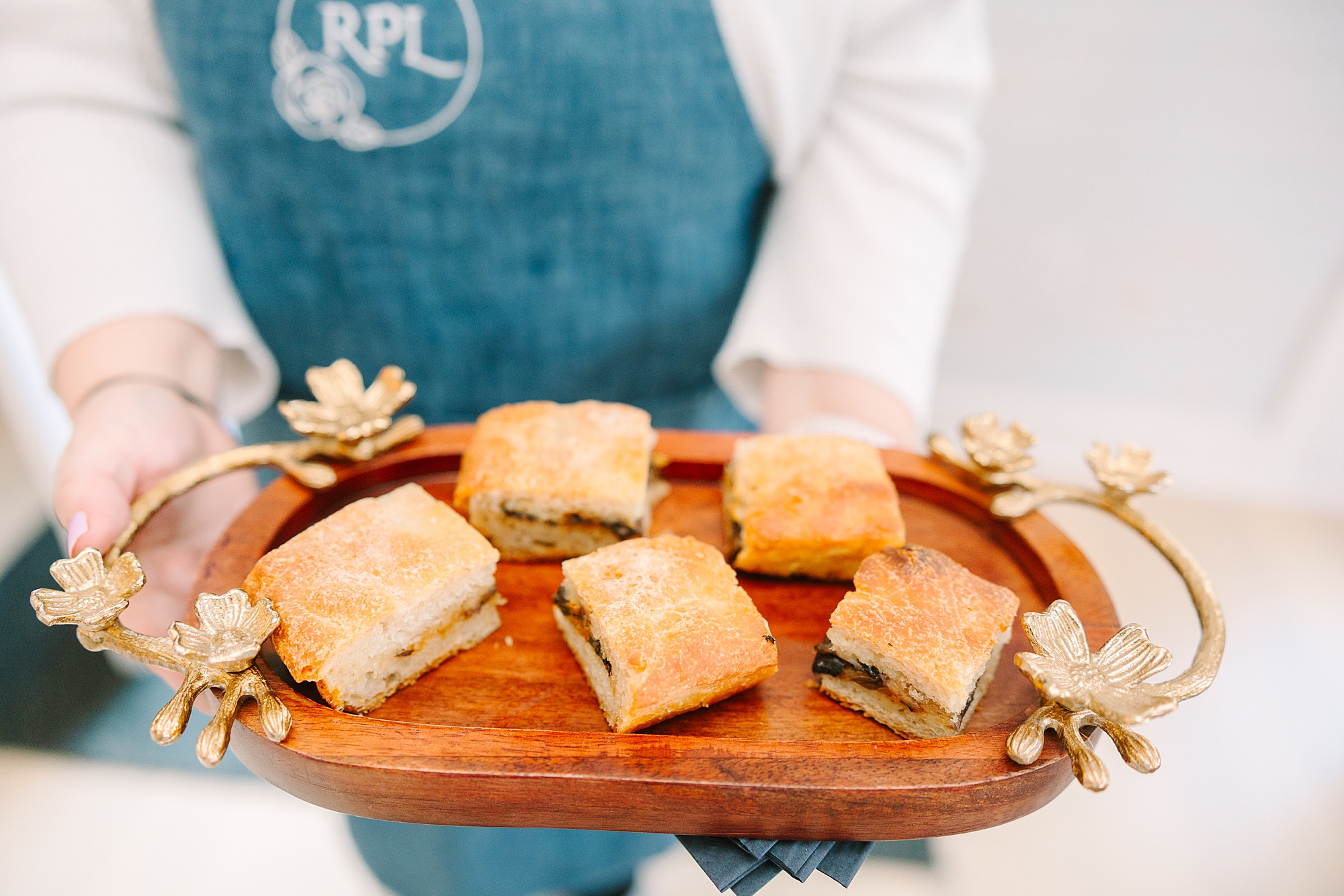 chef in blue apron holds out wooden tray of sandwiches